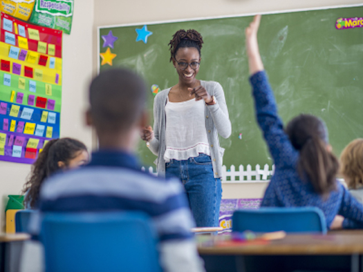 A Black female teacher smiles and points to a student who is raising her hand.