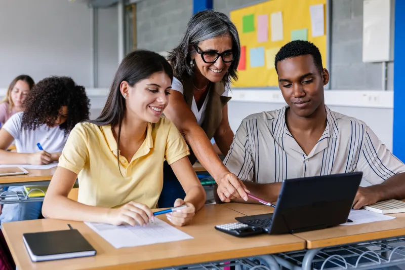 Two high school students sharing a laptop interacting with a teacher 
