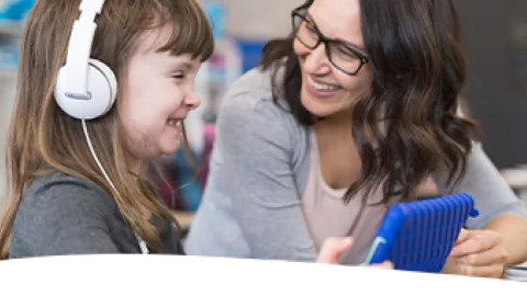 A female elementary student wearing headphones smiles while working with her teacher on a classroom digital tablet.